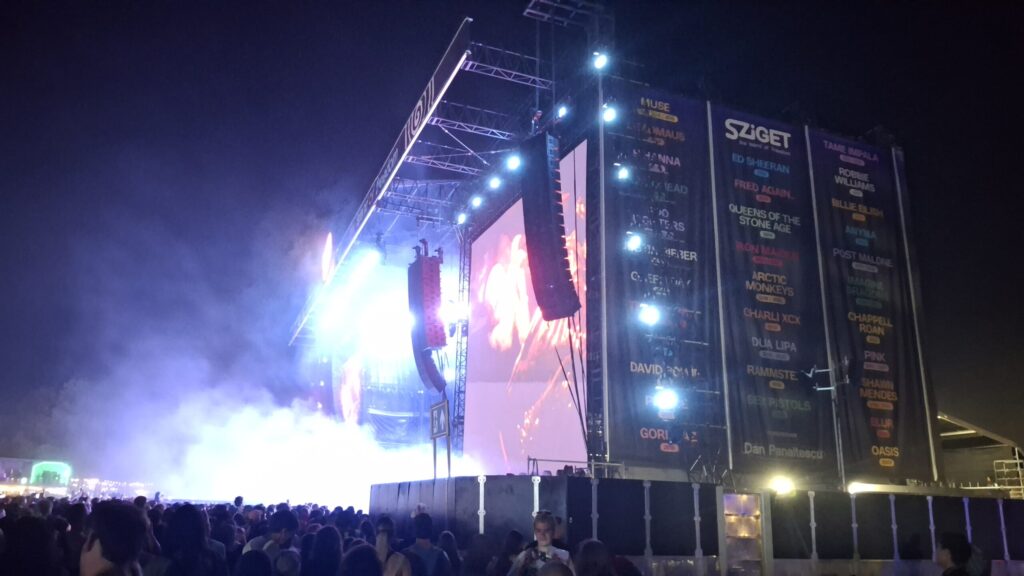 Photo de nuit d'une grande scène du festival Sziget. Des projecteurs et des écrans LED éclairent la scène, tandis qu'une foule dense se rassemble devant celle-ci. Des bannières grand format arborant le logo du Sziget et les noms de divers artistes sont accrochées à la structure latérale de la scène.